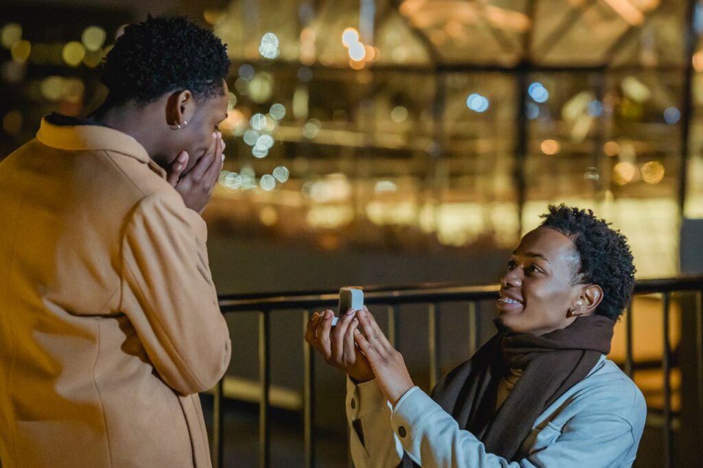 Side view of African American knelling and doing proposal to amazed homosexual lover at night